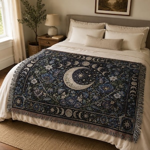 Horizontal woven blanket wall hanging with a large crescent moon, moon phase border, stars, deep blue flowers, white blossoms, dark botanical vines, ornate celestial frame, and fringed textile edges.