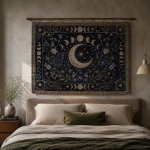 Horizontal woven blanket wall hanging with a large crescent moon, moon phase border, stars, deep blue flowers, white blossoms, dark botanical vines, ornate celestial frame, and fringed textile edges.