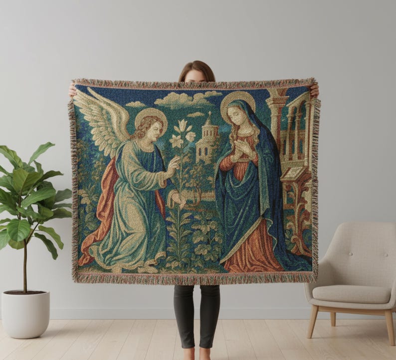 Horizontal woven blanket wall hanging featuring Archangel Gabriel kneeling before the Virgin Mary in an Annunciation scene, with lily flowers, halos, soft robes, garden landscape, old architecture, muted vintage colors, and fringed textile edges.
