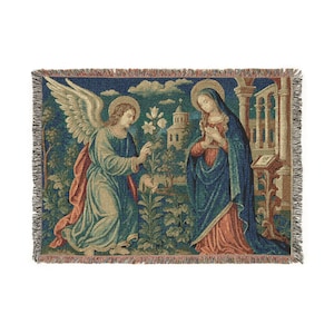 Horizontal woven blanket wall hanging featuring Archangel Gabriel kneeling before the Virgin Mary in an Annunciation scene, with lily flowers, halos, soft robes, garden landscape, old architecture, muted vintage colors, and fringed textile edges.