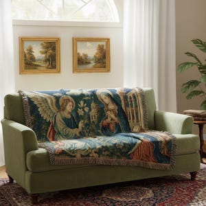 Horizontal woven blanket wall hanging featuring Archangel Gabriel kneeling before the Virgin Mary in an Annunciation scene, with lily flowers, halos, soft robes, garden landscape, old architecture, muted vintage colors, and fringed textile edges.