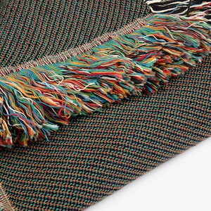 Horizontal woven blanket wall hanging featuring Archangel Gabriel kneeling before the Virgin Mary in an Annunciation scene, with lily flowers, halos, soft robes, garden landscape, old architecture, muted vintage colors, and fringed textile edges.