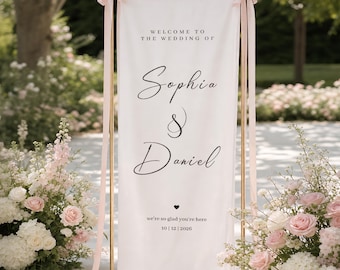 Könnte beinhalten: Ein vertikales Hochzeits-Willkommensschild aus weißem Stoff, das an einem goldfarbenen Metallrahmen hängt und an den oberen Ecken mit hellrosa Bändern befestigt ist. Das Schild trägt den schwarzen Text: WELCOME TO THE WEDDING OF Sophia & Daniel, we're so glad you're here, 10 | 12 | 2026.