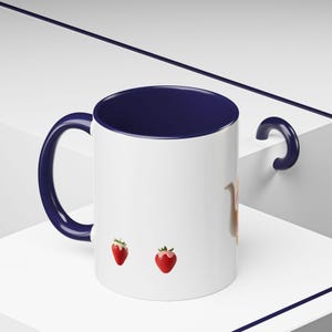 May include: White ceramic mug with a navy blue interior, handle, and rim. The mug features two red strawberries with green leaves. A whimsical design for a coffee or tea cup.