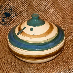 May include: A handmade ceramic bowl with a lid, featuring a spiral pattern in blue, brown, and cream. The lid has a teal knob. The bowl sits on a textured, tan background. The bowl is a decorative item.