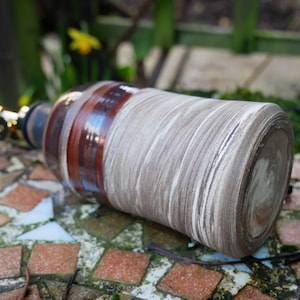 Puede incluir: Una botella de cerámica con boquilla de latón y diseño decorativo. La botella tiene un patrón de remolino marrón claro y blanco con bandas marrones oscuras. La botella está sobre una superficie de mosaico.