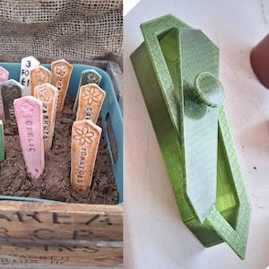 May include: A collection of ceramic garden markers with embossed floral designs and plant names like "Garlic" and "Tomatoes" in a wooden crate. A green plastic tool with a circular knob is also shown.