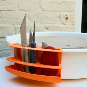 Peut inclure: Un organisateur d'outils en plastique orange vif fixé sur le côté d'un tour de potier blanc. L'organisateur comporte plusieurs fentes contenant divers outils de sculpture en bois et en métal, dont une mirette rouge et une aiguille.