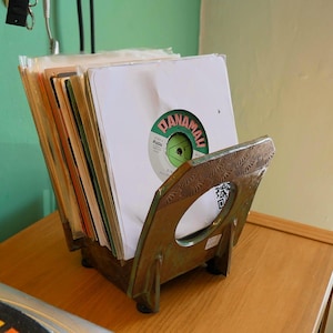 May include: A vintage record holder made of metal, holding several vinyl records. The holder has a decorative design and a circular opening. The records are in white sleeves, with one showing a green and white label.