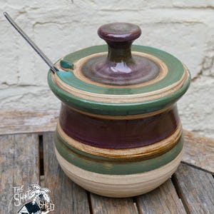 May include: A handmade ceramic sugar bowl with a lid and a small spoon. The bowl is cylindrical with horizontal stripes in shades of green, brown, and purple. The lid has a matching color scheme and a rounded knob. The spoon is silver.
