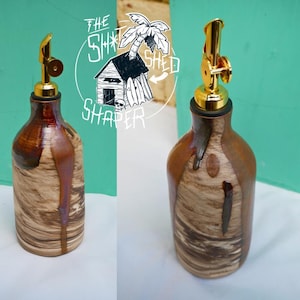 May include: Two decorative oil bottles with gold spouts. The bottles have a wood-grain pattern with brown and tan tones. The text "The Shaka Shed Shaper" is in a circle behind the bottles.