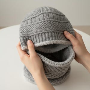 以下が含まれることがあります： グレーのニット帽とスカーフのセット。帽子はリブ編みで、クラウンにテクスチャード加工が施されています。内側は柔らかく明るい色の素材で裏打ちされています。スカーフは帽子に付いており、お揃いのニットパターンになっています。