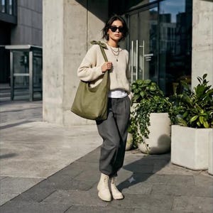 May include: An olive green tote bag with a knotted strap is carried over the shoulder. The bag is paired with a cream-coloured jumper, dark grey trousers, and cream-coloured boots. The person is wearing sunglasses and a necklace.