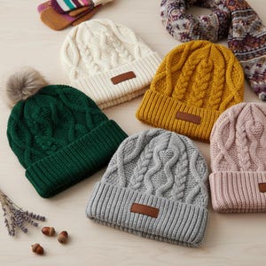 Könnte beinhalten: Eine Auswahl an Zopfstrick-Mützen in Creme, Senfgelb, Waldgrün, Grau und Rosa. Jede Mütze hat einen umgeschlagenen Saum und ein kleines braunes Kunstlederetikett. Eine grüne Mütze ist mit einem Kunstfell-Bommel versehen.