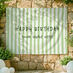 May include: A green and white striped banner with the text "HAPPY BIRTHDAY your Name Here" in dark green lettering. The banner is hanging on a stone wall, with plants and decorative spheres in the foreground.