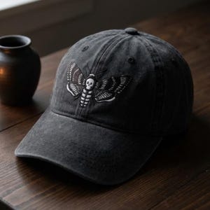 May include: A dark gray baseball cap featuring an embroidered moth design with a skull on the front. The moth is white with black detailing. The cap has a curved brim and a button on top.