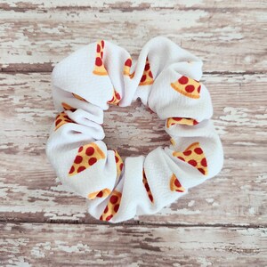 May include: A white fabric hair scrunchie featuring a repeating pattern of pizza slices with yellow crusts, red sauce, and brown pepperoni toppings. The accessory has a gathered, ruffled texture throughout the circular band design.