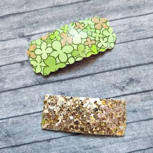 May include: Two hair clips, one with a green floral pattern and one with gold glitter.