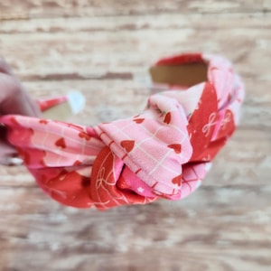 May include: A knotted headband featuring a patchwork design with red and pink fabrics. The fabrics are patterned with hearts and bows. The headband is a Valentine's Day accessory.