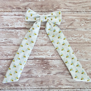 May include: A white fabric bow with a bee pattern. The bow is tied with long tails that have the same bee pattern.