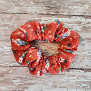 May include: A red scrunchie with a repeating pattern of nutcrackers, Christmas trees, and stars. The fabric is a vibrant red, and the design includes white, blue, and green accents. This hair accessory is perfect for the holiday season.