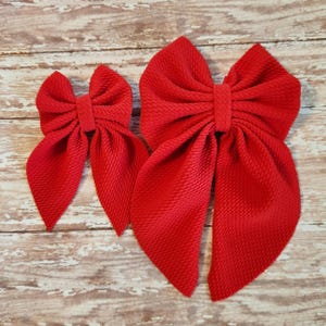 May include: Two red fabric hair bows, one large and one small, with a textured surface. The bows are tied in a classic bow shape.