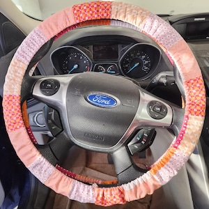 May include: A colorful, patchwork steering wheel cover in shades of pink, orange, and white. The cover is made of fabric and is designed to fit over a car's steering wheel. The Ford logo is visible in the center of the steering wheel.