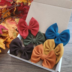 May include: A set of six fabric hair bows in a cardboard box, featuring textured material in solid colors including rust red, olive green, dark brown, teal blue, mustard yellow, and burnt orange, arranged in a cluster.