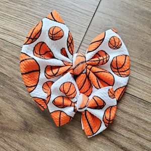 May include: Two white hair bows with a basketball pattern. The bows are made of a soft, textured fabric and feature orange and black basketballs.