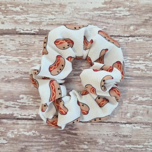 May include: A white fabric hair scrunchie featuring a repeating pattern of small, cartoon-style hot dogs with brown buns, tan sausages, and yellow mustard lines. The accessory has a gathered, ruffled texture throughout its circular shape.