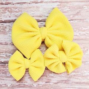 May include: Three yellow fabric hair bows of different sizes. The bows are made of a textured fabric and are arranged in a pyramid shape.