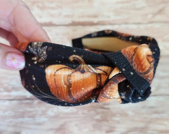 May include: A knotted fabric headband featuring a black background with an orange pumpkin print and small white speckles. The design includes detailed illustrations of pumpkins and foliage, with a central knot detail on the top of the band.