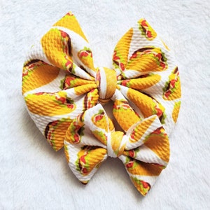 May include: A large, handmade hair bow featuring a repeating pattern of yellow and red tacos on a white background. The bow is tied in the center with a matching fabric strip. The bow is a fun accessory for any occasion.