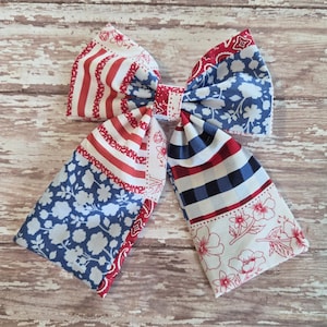 May include: A large fabric bow featuring a patchwork design with red, white, and blue patterns. The bow includes stripes, floral prints, and a plaid pattern, all in patriotic colors. The bow is likely for hair or as a decorative accessory.