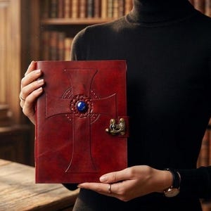 May include: A red leather journal with a cross design and a blue gemstone in the center. The journal has a clasp closure. The cover is embossed with a cross design. The journal is held in front of a bookshelf.