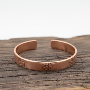 Puede incluir: Una pulsera de brazalete de color cobre con un diseño abierto. La pulsera presenta el símbolo Om grabado en la parte delantera. La pulsera se muestra sobre una superficie de madera envejecida.