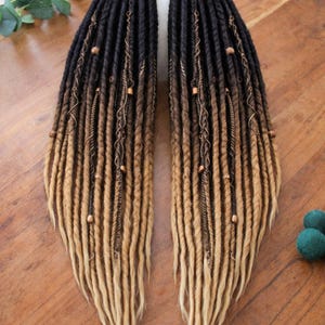 May include: Two sets of dreadlocks with a gradient effect, transitioning from black at the roots to brown and then blonde at the tips. The dreads are adorned with copper-colored beads and decorative wire wraps. The hair extensions are displayed on a wooden surface.