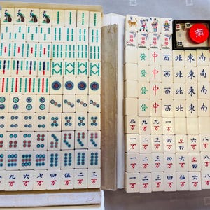 Puede incluir: Un juego de Mahjong vintage con fichas de color marfil, con varios símbolos y caracteres en rojo, verde y azul. El juego incluye dados y un marcador rojo, todo guardado en una caja de cartón.
