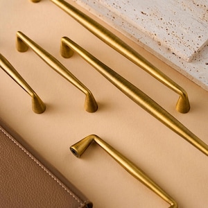 May include: A selection of gold-coloured cabinet pulls and handles in different sizes. The hardware has a brushed finish and a minimalist design. The pulls are arranged on a neutral background, with a leather-bound notebook and a stone tile in the background.