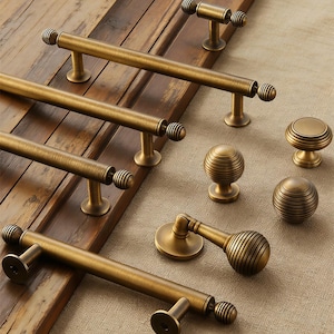 May include: A selection of antique brass cabinet hardware, including long bar pulls, round knobs, and a curved handle. The hardware has a warm, aged finish and ribbed detailing. The pieces are arranged on a wooden door and neutral fabric.