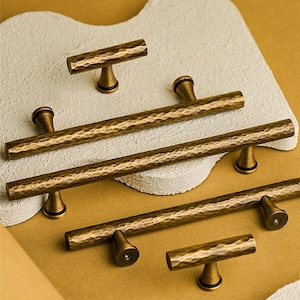 May include: A selection of antique brass cabinet handles, featuring T-bar and long bar pulls. The hardware has a hammered texture and a warm, aged finish. The pulls are displayed on a textured, off-white surface against a golden background.