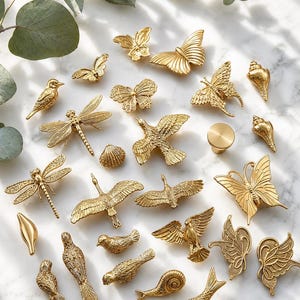 Puede incluir: Una colección de tiradores y pomos decorativos dorados en varias formas, incluyendo mariposas, pájaros, libélulas y conchas marinas. La ferretería está dispuesta sobre una superficie similar al mármol.