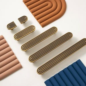 May include: An assortment of brass-toned cabinet hardware, including knobs and pulls, arranged with terracotta and blue silicone trivets. The hardware features a grooved design. The trivets are in a curved, arched shape.