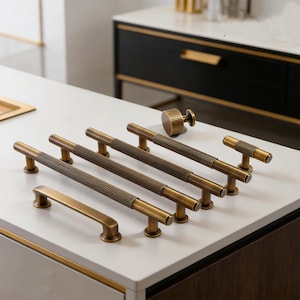May include: A selection of antique brass cabinet handles and knobs. The collection includes ribbed bar pulls, a round knob, and a T-shaped pull. The hardware is displayed on a white surface, highlighting the different designs.