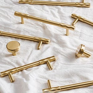 May include: A selection of gold-toned cabinet handles and knobs. The hardware has a geometric diamond pattern on the handles and knobs. The items are displayed on a white, textured surface.