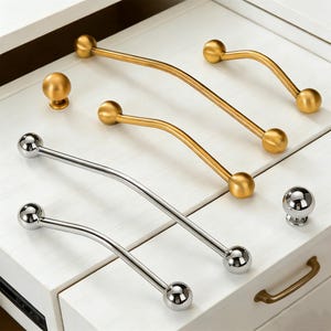 May include: A selection of cabinet hardware, including gold and silver-toned curved handles and round knobs. The handles have a ball-shaped design on each end. The hardware is displayed on a white cabinet.