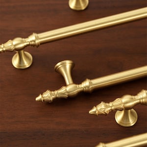 May include: Close-up of several antique brass cabinet pulls and handles. The hardware features ornate detailing and a warm, golden finish. The pulls are arranged on a dark wood surface, showcasing their design and texture.