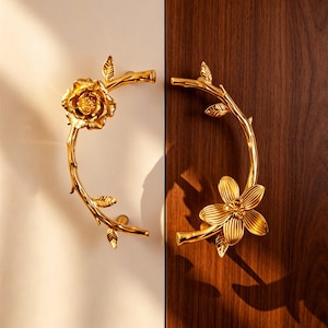 May include: Two gold-coloured door handles, each shaped like a floral branch. One handle features a rose, the other a flower with multiple petals. The handles are mounted on a white and a dark brown wooden surface.