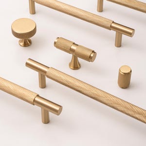 Peut inclure: Une collection de quincaillerie de meubles dorée, comprenant des barres de tirage, des boutons et une poignée en forme de T. La quincaillerie présente un motif texturé en losanges. Les articles sont disposés sur une surface blanche, mettant en valeur leurs formes et tailles.