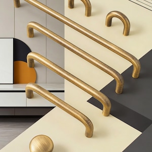 May include: A selection of antique brass cabinet hardware, featuring bar pulls and a round knob. The pulls are of varying sizes, with a curved design. The hardware has a textured finish, arranged on a geometric background.